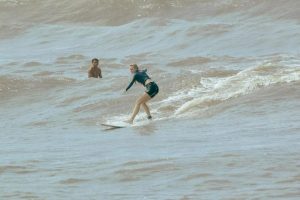 A woman wearing a dark wetsuit is standing on a white surfboard, gliding over the waves