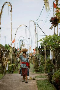 An elderly woman walks along a path in a village in Bali, carrying a woven basket on her head