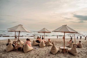 Relaxed atmosphere by the sandy beach at sunset with several people sitting on bean bags under large umbrellas