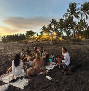 A group of people are gathering on a dark sandy beach at sunset