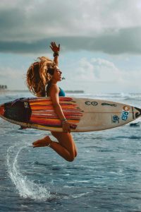 A young female surfer jumping joyfully while holding her surfboard at the water's edge