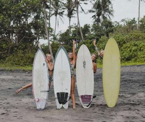 Four smiling women standing on a black sand beach with their surfboards and palm trees in the background