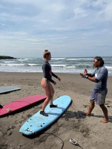 A person practicing surf stances on a blue soft-top board under the guidance of an instructor on a sandy beach
