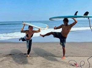 Two individuals holding surfboards above their heads on a sandy beach with blue ocean water in the background
