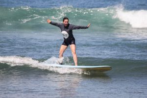 a beginner surfer practicing balancing on a longboard while wearing a dark rash guard and board shorts during a surf lesson