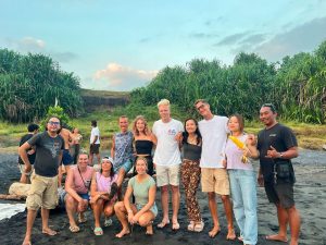 a group of people posing together on a dark sand beach with lush green vegetation in the background