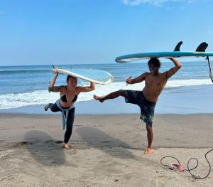 two people balancing on one leg on a sandy beach while holding blue surfboards above their heads