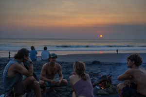 a group of friends relaxing around a small bonfire on a sandy beach during sunset, with waves rolling in the background