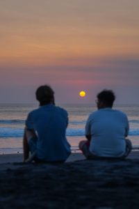 two people sitting on a sandy beach and watching the sunset over the ocean