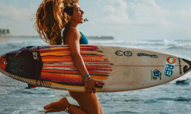 A young female surfer jumping joyfully while holding her surfboard at the water's edge