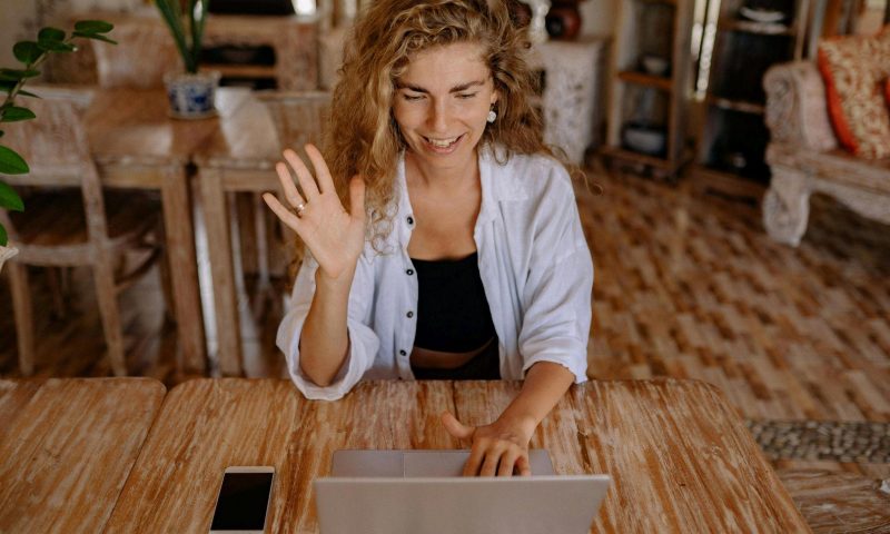 A young woman is making a video call using her laptop at a wooden table
