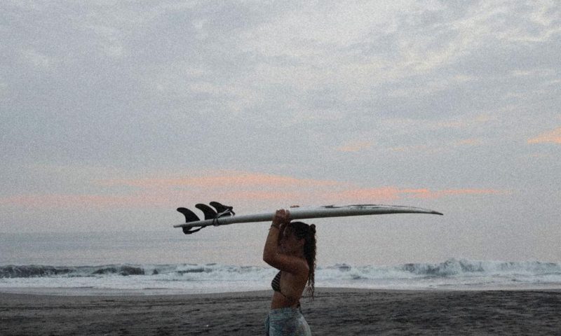 Surfer holding a surfboard overhead on a black sand beach in Bali at dusk
