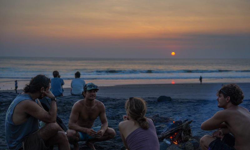 a group of friends relaxing around a small bonfire on a sandy beach during sunset, with waves rolling in the background