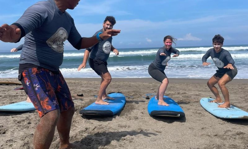 a surf instructor leading a beginner pop-up technique lesson on the sandy beach with students practicing on soft-top boards